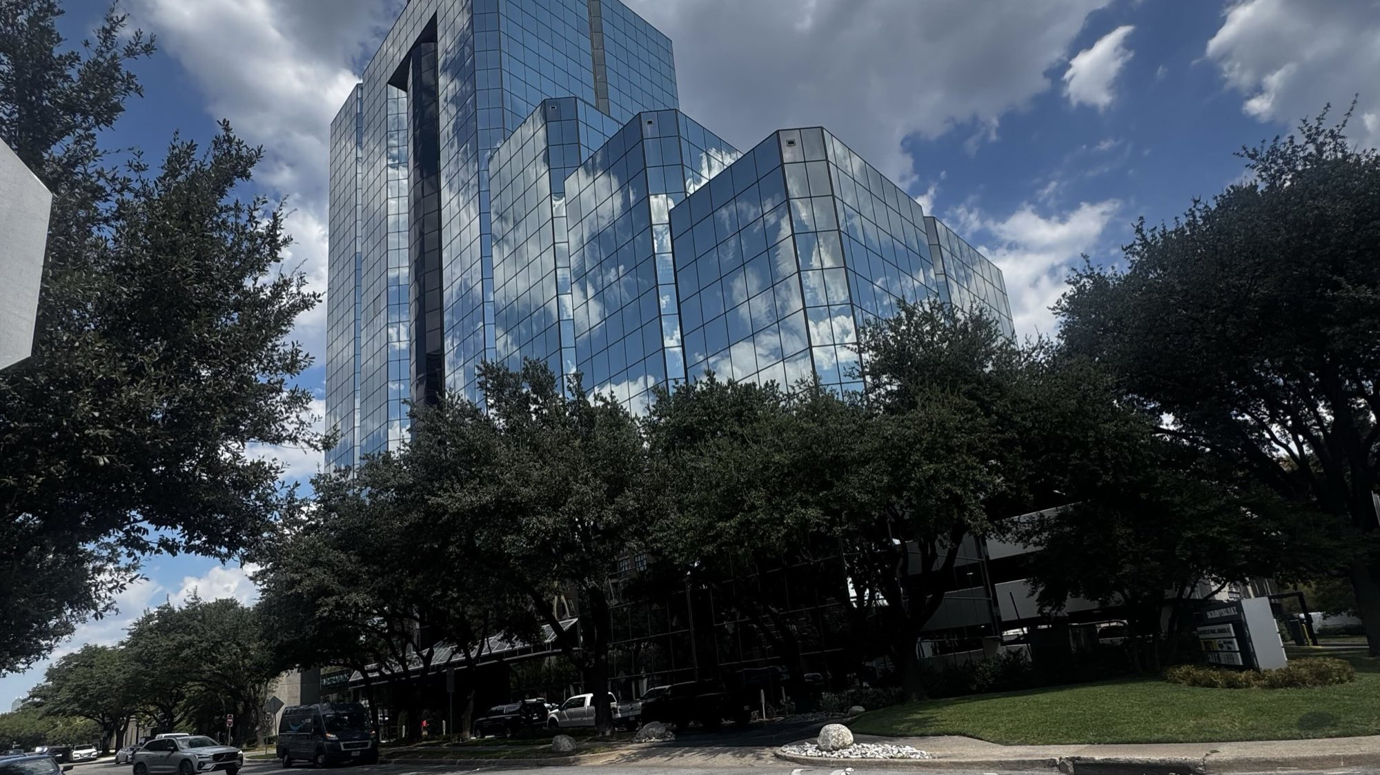 Highland Park Place, the mirrored glass office tower at 4514 Cole Avenue in Dallas, seen from the sidewalk through mature live oak branches